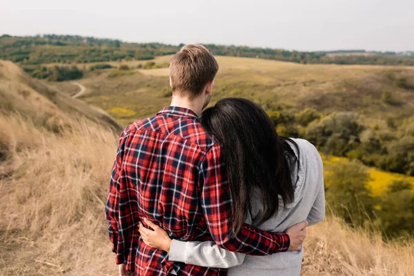 Rückansicht eines multiethnischen Paares, das sich mit grasbewachsener Landschaft auf verschwommenem Hintergrund umarmt — Stockfoto