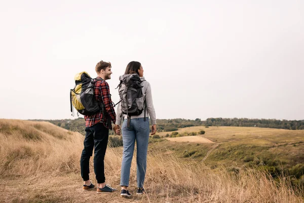 Pareja sonriente de excursionistas multiétnicos tomados de la mano mientras miran el paisaje sobre un fondo borroso - foto de stock