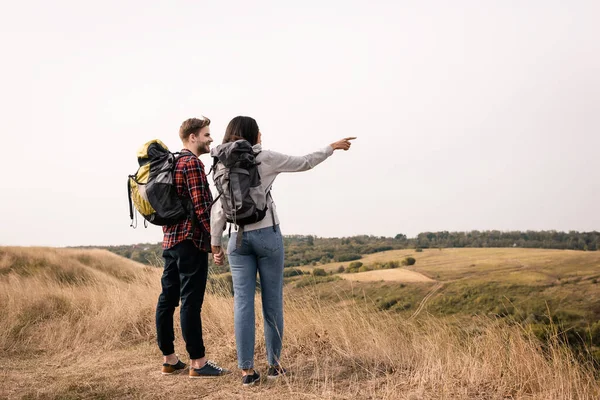 Smiling hiker holding hand of african american girlfriend pointing with finger with grassy hills on blurred background — Stock Photo