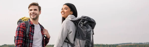 Senderistas multiétnicos sonrientes con mochilas mirando al aire libre, pancarta - foto de stock