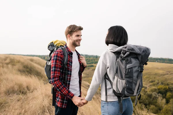 Lächelnder Mann hält Hand seiner afrikanisch-amerikanischen Freundin mit Rucksack auf Rasen — Stockfoto