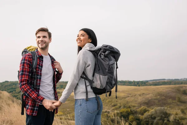 Sorridente coppia multietnica con zaini che si tengono per mano con colline su sfondo sfocato — Foto stock