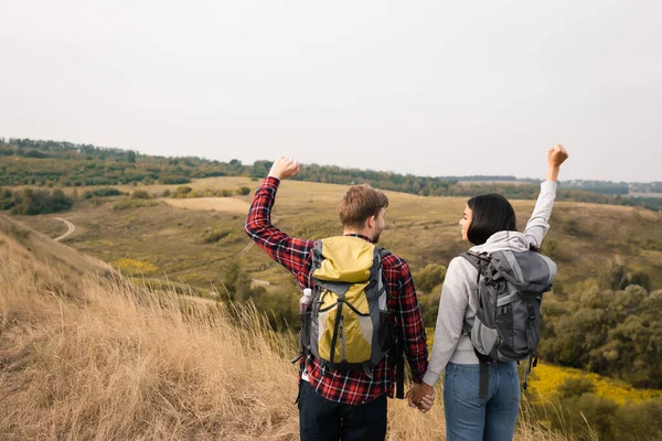 Smiling african american woman with backpack showing yeah gesture and holding hand of boyfriend with hills on blurred background — Stock Photo