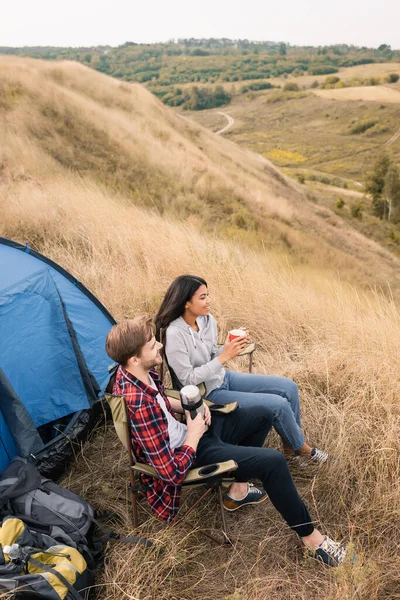 Высокий угол зрения улыбающихся мультиэтнических туристов с чашкой и термосом сидит рядом рюкзаки и палатки на газоне — стоковое фото