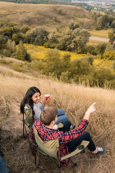 Overhead Ansicht des Mannes zeigt mit dem Finger in der Nähe lächelnde afrikanisch-amerikanische Freundin mit Tasse auf Stuhl auf dem Feld — Stockfoto