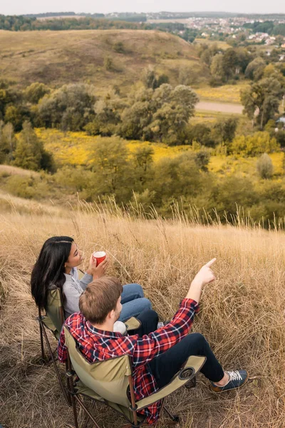 Вид зверху чоловіка, що вказує пальцем біля усміхненої афроамериканської жінки з чашкою під час кемпінгу на газоні — стокове фото