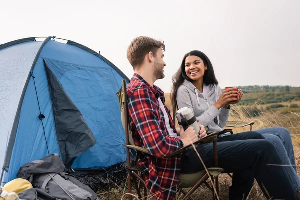 Smiling interracial couple smiling while holding cup and thermos near tent on lawn — Stock Photo