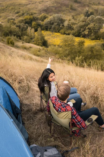 Vue aérienne de la femme afro-américaine souriante avec tasse pointant du doigt près du petit ami et de la tente sur la pelouse — Photo de stock