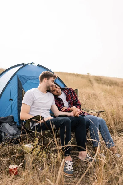 Улыбающаяся многонациональная пара, держащаяся за руки, сидя возле палатки на газоне — стоковое фото