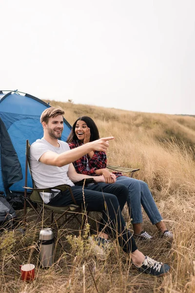 Smiling man pointing with finger while holding hand of african american girlfriend during camping on lawn — Stock Photo