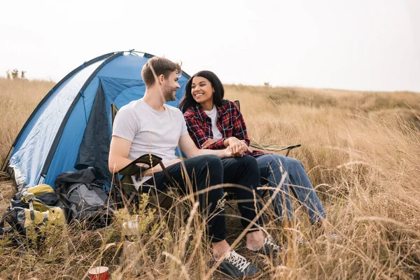 Мультиэтнические путешественники держатся за руки, улыбаясь друг другу возле палатки на газоне — стоковое фото
