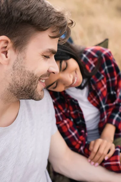 Sonriente hombre con los ojos cerrados cerca de la novia afroamericana sobre fondo borroso durante el viaje - foto de stock