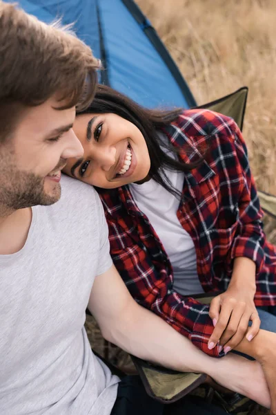 Sonriente mujer afroamericana mirando al novio en primer plano borroso durante el campamento en el césped - foto de stock