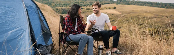 Sorridente coppia multietnica con tazza e thermos parlando vicino tenda durante il campeggio, banner — Foto stock