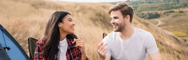 Hombre alegre hablando con la novia afroamericana en sillas durante el camping en el césped, pancarta - foto de stock