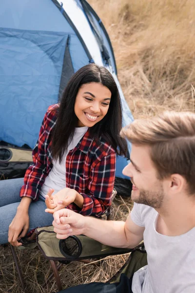 Улыбающаяся африканская американка смотрит на своего парня на размытом переднем плане возле палатки во время похода — стоковое фото