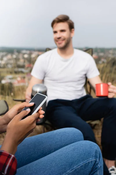 Mulher afro-americana usando smartphone perto de namorado sorridente com copo e garrafa térmica em primeiro plano borrado durante o acampamento — Fotografia de Stock