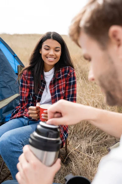 Sonriente mujer afroamericana sosteniendo taza cerca de novio con termos en primer plano borroso durante el camping - foto de stock