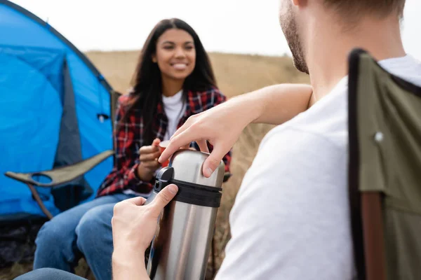 Hombre sosteniendo termos cerca de la sonriente mujer afroamericana sobre fondo borroso durante el campamento en el prado - foto de stock