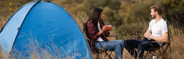 Sonriente pareja interracial con taza sentada en sillas cerca de la tienda durante el campamento, pancarta — Stock Photo