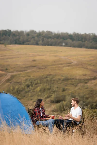 Uomo che parla con sorridente fidanzata afro-americana con coppa durante il campeggio vicino tenda con campo su sfondo sfocato — Foto stock