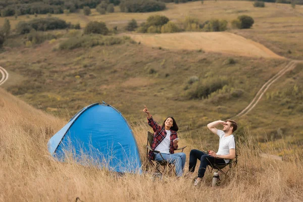 Lächelnde Afroamerikanerin hält Smartphone in der Hand und zeigt mit dem Finger auf Freund mit Tasse und Zelt auf Rasen — Stockfoto