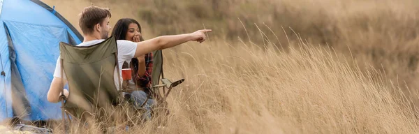 Uomo che punta con il dito vicino alla ragazza afro-americana con coppa durante il campeggio sul prato, banner — Foto stock