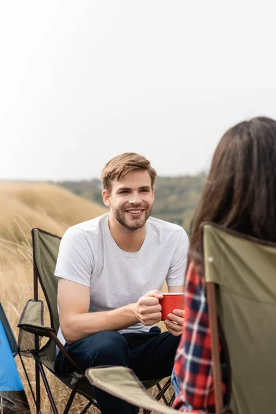 Uomo sorridente con coppa guardando la ragazza sulla sedia in primo piano sfocato durante il campeggio — Foto stock