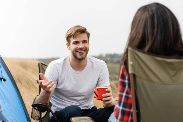 Sonriente hombre con taza hablando con su novia en primer plano borroso durante el camping - foto de stock