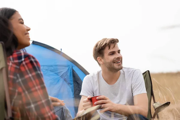 Smiling man holding cup while sitting near african american girlfriend on blurred foreground and tent on lawn — Stock Photo