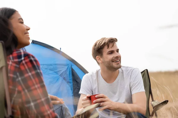 Uomo sorridente che tiene la tazza mentre si siede vicino alla ragazza americana africana in primo piano offuscata e tenda sul prato — Foto stock