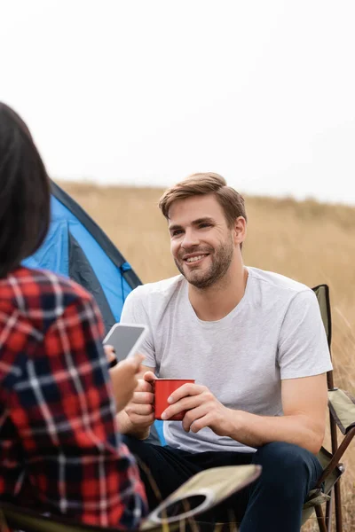 Hombre sonriente sosteniendo taza cerca de novia afroamericana con teléfono inteligente en primer plano borroso durante el camping - foto de stock