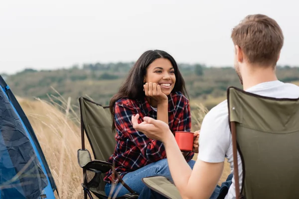 Femme afro-américaine souriante tenant une tasse et regardant son petit ami au premier plan flou pendant le camping — Photo de stock