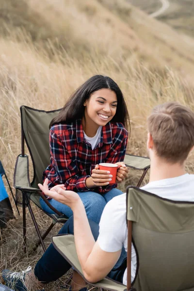 Positiva mujer afroamericana con taza sentada cerca de novio durante acampar en campo herboso - foto de stock