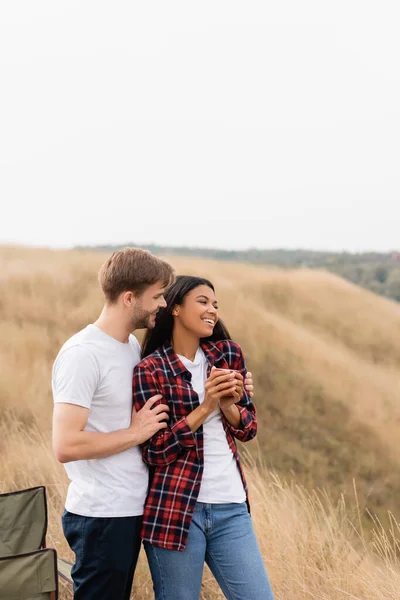 Homme souriant étreignant petite amie afro-américaine avec tasse sur prairie herbeuse — Photo de stock