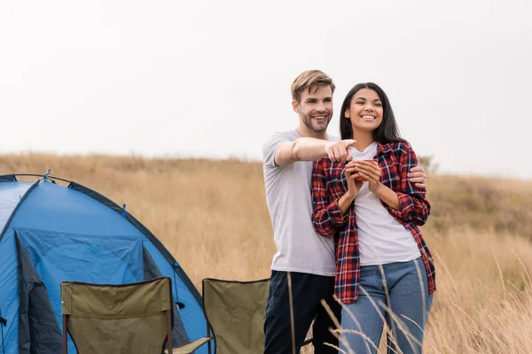 Homme souriant embrassant petite amie afro-américaine avec tasse et pointant du doigt pendant le camping sur la pelouse — Photo de stock