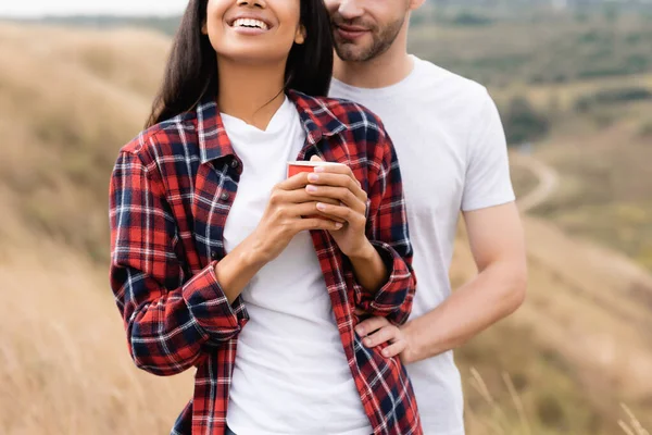 Vista recortada del hombre abrazando sonriente mujer afroamericana con taza al aire libre - foto de stock