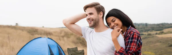 Mujer afroamericana sonriendo mientras abraza novio con tienda de campaña sobre fondo borroso en el prado, bandera - foto de stock