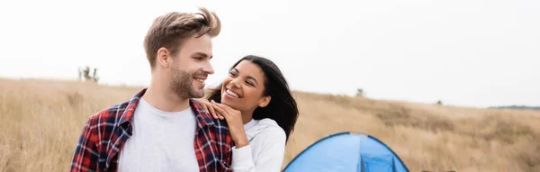 Smiling man looking at african american girlfriend with tent and field on blurred background, banner — Stock Photo