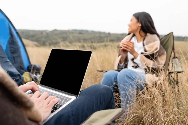 Uomo utilizzando laptop vicino fidanzata afro-americana con coppa su sfondo sfocato durante il campeggio in campo — Foto stock