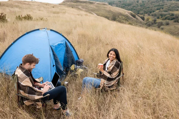 Sorridente donna africana americana in possesso di tazza vicino fidanzato con computer portatile e tenda sul prato — Foto stock