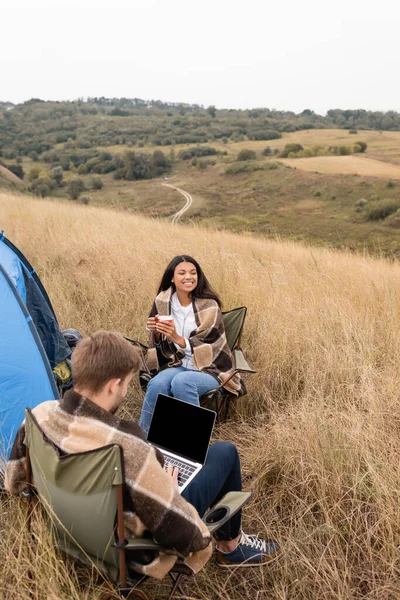 Sorridente donna afro-americana con coppa seduta vicino al fidanzato con laptop in primo piano sfocato e tenda all'aperto — Foto stock
