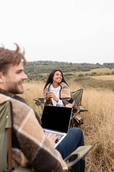 African american woman wrapped in blanket holding cup near boyfriend with laptop on chairs on lawn — Stock Photo