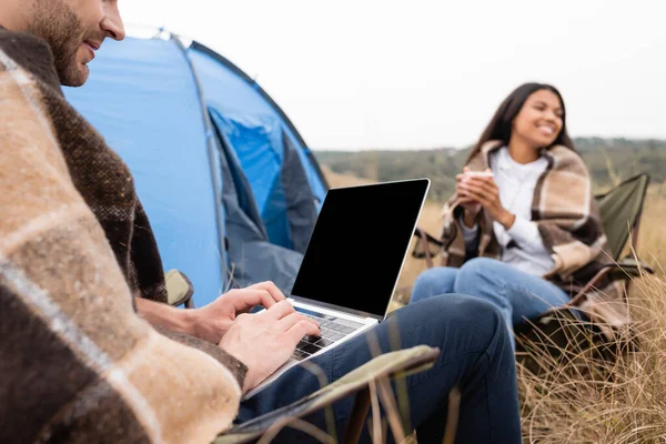 Hombre usando el ordenador portátil cerca sonriente mujer afroamericana con taza durante el camping sobre fondo arrugado - foto de stock