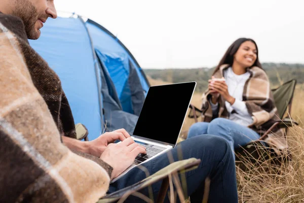 Mann benutzt Laptop in der Nähe lächelnde Afroamerikanerin mit Tasse beim Zelten auf grundiertem Hintergrund — Stockfoto