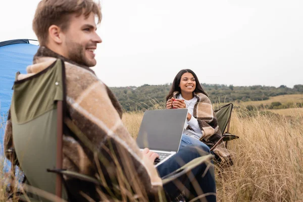 Cheerful african american woman in blanket holding cup near boyfriend with laptop on blurred foreground during camping — Stock Photo