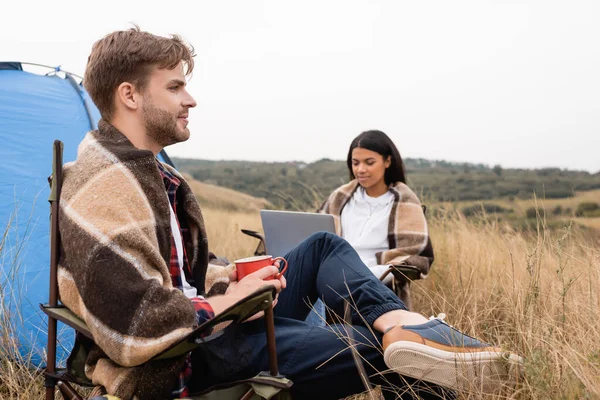 Man wrapped in blanket holding cup near african american girlfriend using laptop on lawn on blurred background — Stock Photo