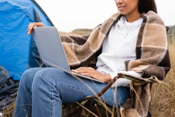 Vue recadrée de la femme afro-américaine enveloppée dans une couverture à l'aide d'un ordinateur portable pendant le camping — Photo de stock