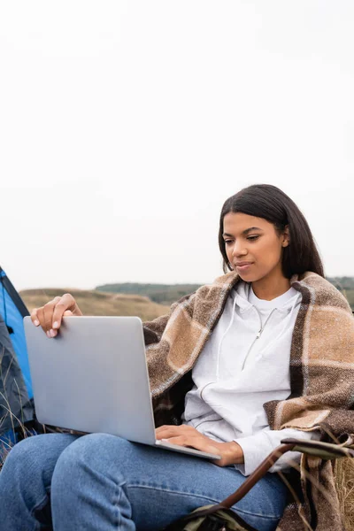 Africano americano mulher envolto em cobertor usando laptop durante o acampamento ao ar livre — Fotografia de Stock