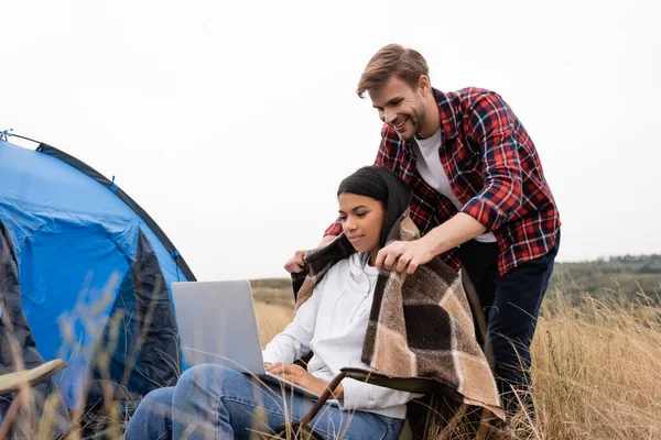 Femme afro-américaine utilisant un ordinateur portable près copain souriant avec couverture pendant le camping sur prairie — Photo de stock